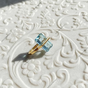 Könnte beinhalten: Ein goldener Ring mit zwei rechteckigen, hellblauen Edelsteinen. Die Edelsteine sind nebeneinander angeordnet, mit einem goldenen Balken oben. Der Ring liegt auf einer weißen, floral gemusterten Oberfläche. Die Worte "ENDLESS EMERALD" sind gedruckt.
