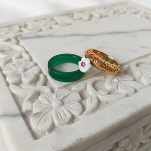 May include: Two rings sit on a white, floral-carved surface. One is a vivid green band, decorated with a white flower and a pink centre. The other is a gold ring with an intricate, swirling pattern. Jewellery.