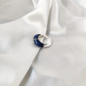 Könnte beinhalten: Ein silberner Ring mit einem blauen Lapislazuli-Stein, der in die Band eingefasst ist. Der Ring liegt auf einem weißen Satin-Hintergrund.