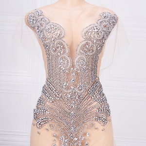 May include: A sheer, nude-colored bodysuit with a decorative design made of silver rhinestones. The design features a V-neckline and intricate patterns.