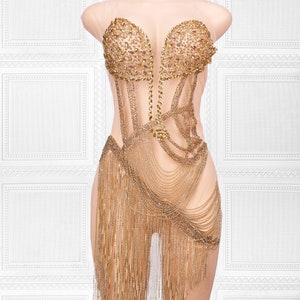 Könnte beinhalten: Ein durchsichtiges goldenes Kleid mit einem perlenbesetzten Oberteil und einem Fransenrock. Das Kleid ist mit Strasssteinen und Pailletten verziert.