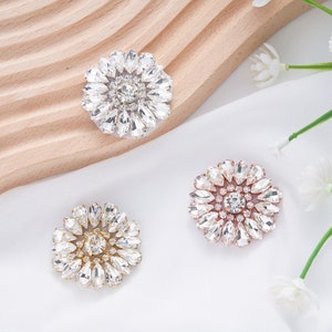 May include: Three decorative flower-shaped embellishments with crystal rhinestones. The embellishments are in silver, gold, and rose gold.