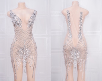 May include: A sheer, sleeveless dress with a plunging neckline and a beaded design. The dress is covered in silver rhinestones and has a flowing skirt.