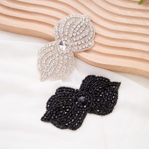 Può includere: Due abbellimenti decorativi a forma di fiocco. Uno è argentato con strass trasparenti e un cristallo centrale. L'altro è nero con perline nere e una gemma nera centrale. Entrambi sono su una superficie bianca con un accento di legno.