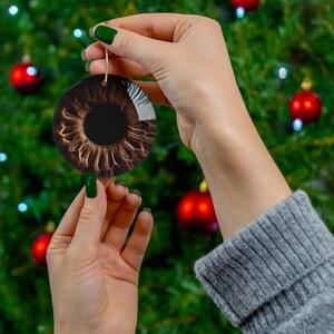 May include: A round Christmas ornament with a brown eye printed on it. The ornament is being held by a hand with green nail polish.