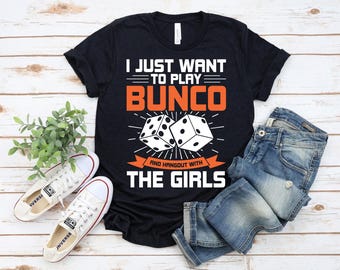 Op de afbeelding: Zwart T-shirt met de tekst "I JUST WANT TO PLAY BUNCO AND HANGOUT WITH THE GIRLS" in wit en oranje. Het shirt heeft een afbeelding van twee dobbelstenen. Witte sneakers en een blauwe spijkerbroek staan ook op de foto.