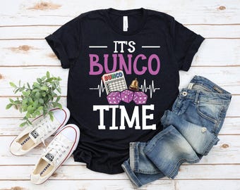 Könnte beinhalten: Ein schwarzes T-Shirt mit den Worten "IT'S BUNCO TIME" in Weiß und Rosa. Das Design umfasst Würfel, eine Bingo-Karte und eine Glocke. Weiße Leinenschuhe und blaue Jeans sind ebenfalls im Bild.