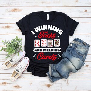 Pode incluir: T-shirt preto com o texto "I Winning Tricks and Melding Cards" em branco e vermelho. A camisa apresenta gráficos de cartas de baralho. Um par de tênis brancos e jeans azuis estão ao lado da camisa.