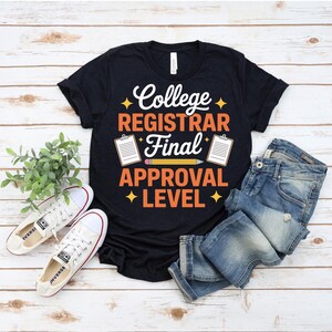 Puede incluir: Camiseta negra con el texto "College Registrar Final Approval Level" en blanco y naranja. La camiseta se combina con vaqueros azules y zapatillas blancas. El diseño incluye un lápiz y portapapeles.