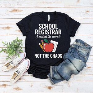 Op de afbeelding: Zwart T-shirt met de tekst "SCHOOL REGISTRAR I control the records NOT THE CHAOS". Het shirt heeft een rode appel, potlood en notitieblok. Witte sneakers en een blauwe spijkerbroek staan ook op de foto.