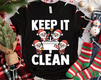 Pode incluir: Camiseta preta com as palavras "KEEP IT CLEAN" em letras brancas. A camiseta apresenta um gráfico de quatro Papais Noéis trabalhando em uma pia com ferramentas. Tema natalino festivo.