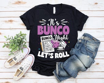 May include: Black t-shirt with the text "It's Bunco Night, Let's Roll" in white and purple. The design includes dice, a score sheet, and a bell. A pair of white sneakers and blue jeans are also in the image.