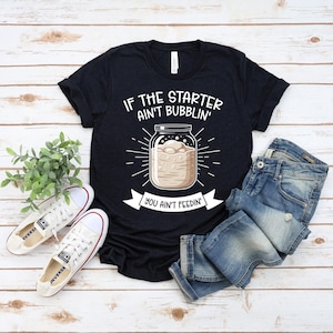 Peut inclure: T-shirt noir avec le texte "IF THE STARTER AIN'T BUBBLIN' YOU AIN'T FEEDIN'". Le graphisme représente un bocal avec une substance pétillante. Des baskets blanches et un jean bleu sont également dans l'image.