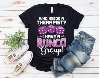 Könnte beinhalten: Schwarzes T-Shirt mit dem Text "WHO NEEDS A THERAPIST? I HAVE A BUNCO GROUP!" und lila Würfelgrafiken. Das Bild zeigt blaue Jeans und weiße Turnschuhe, was einen lässigen Stil suggeriert.