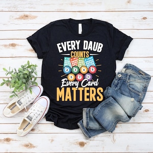 Può includere: T-shirt nero con la scritta "EVERY DAUB COUNTS EVERY CARD MATTERS" e grafica a tema bingo. Il design include palline e cartelle del bingo. L'immagine mostra anche un paio di scarpe da ginnastica bianche e jeans blu.