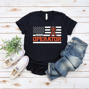 Peut inclure: T-shirt noir avec un motif de drapeau américain et le texte orange "HAM OPERATOR". Le motif du drapeau comprend des étoiles et des rayures, avec un graphique de tour radio. Le t-shirt est associé à un jean bleu et des baskets blanches.