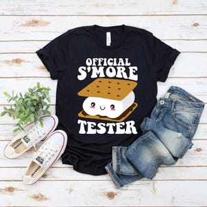 May include: A black t-shirt with the words "OFFICIAL S'MORE TESTER" in white. The graphic features a smiling s'more with a graham cracker, marshmallow, and chocolate. White trainers and blue jeans are also in the image.