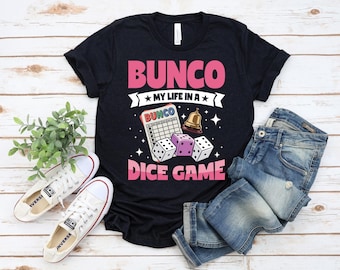 May include: Black t-shirt with the text "Bunco Dice Game" in pink and white. The design includes dice, a bell, and a Bunco score card. The shirt is paired with white sneakers and blue jeans.