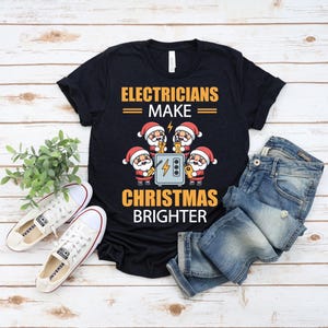 May include: Black t-shirt with the text "Electricians Make Christmas Brighter" in gold. The graphic features cartoon Santas working on an electrical box. White sneakers and blue jeans are also in the image.