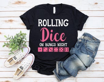 May include: A black t-shirt with the words "ROLLING Dice ON BUNCO NIGHT" in white and pink. Below the text are pink dice illustrations. The shirt is paired with blue jeans and white sneakers.
