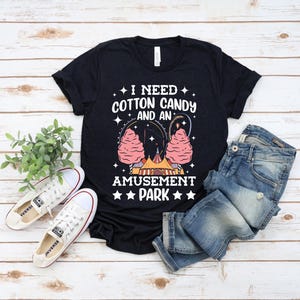 May include: Black t-shirt with the text "I Need Cotton Candy and an Amusement Park" in white. The graphic includes pink cotton candy, a Ferris wheel, and a circus tent. White sneakers and blue jeans are also in the image.