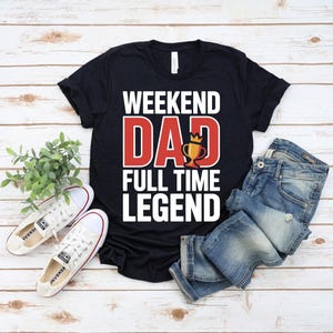 以下が含まれることがあります： 黒の半袖Tシャツ。白い文字で「WEEKEND」、赤い文字で「DAD」（Aの代わりに金のトロフィーのアイコン）、白い文字で「FULL TIME LEGEND」とプリントされている。青いデニムパンツと白いスニーカーが添えられている。