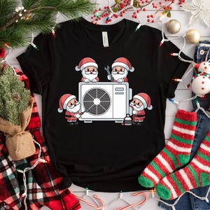 May include: Black t-shirt featuring a cartoon design of Santa Claus characters repairing an air conditioning unit. The Santas are wearing red suits and hats. The air conditioner is white with a circular fan.