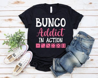 May include: Black t-shirt with the text "BUNGO Addict IN ACTION" in white and pink lettering. Below the text are pink dice illustrations. The shirt is paired with blue jeans and white trainers.