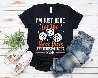 May include: Black t-shirt with white and orange text that reads "I'M JUST HERE for the Three Dices SAID NO BUNCO PLAYER EVER". The shirt features three dice illustrations. A pair of white sneakers and blue jeans are also in the image.