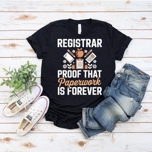 May include: Black t-shirt with the text "REGISTRAR PROOF THAT PAPERWORK IS FOREVER" in white and orange. The shirt features graphic elements of a pencil, apple, and notepad. White sneakers and blue jeans are also in the image.