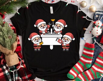 Pode incluir: Camiseta preta com uma ilustração de desenho animado de quatro Papais Noéis usando ferramentas em uma pia. Os Papais Noéis usam trajes e chapéus vermelhos. Um design divertido e festivo para a época natalícia.