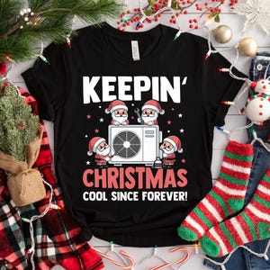 May include: Black t-shirt with the text "KEEPIN' CHRISTMAS COOL SINCE FOREVER!" and a graphic of Santa Claus characters around an air conditioning unit. The shirt is surrounded by Christmas decorations.