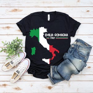 May include: A black t-shirt featuring a map of the Emilia-Romagna region of Italy in green, white, and red. The text "EMILIA-ROMAGNA ITALY" is printed on the shirt. The shirt is paired with blue jeans and white sneakers.