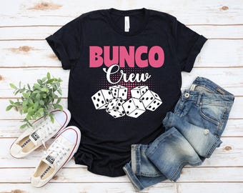 May include: A black t-shirt with the text "BUNCO Crew" in pink and white, with dice illustrations. The image includes white trainers and blue jeans, suggesting a casual, game-themed outfit.