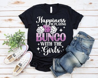 Op de afbeelding: Zwart T-shirt met de tekst "Happiness is Playing Bunco with the Girls" in wit en paars. Het ontwerp bevat dobbelstenen. De afbeelding toont ook witte sneakers en een blauwe jeans.