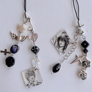 May include: Two phone charms featuring black cords and silver-tone chains. The left charm includes a square acrylic pendant with a male anime character, a cross, wings, a die, and black beads. The right charm features a diamond-shaped pendant with an anime character, a star, a cupcake, an evil eye bead, and a letter L charm.