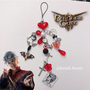 May include: A red and black charm bracelet with a heart, a key, a yin yang symbol, a ghost, a butterfly, a star, a cross, and a portrait of a man. The bracelet is attached to a phone charm.
