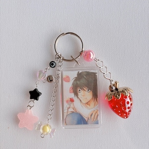 May include: Keychain featuring a rectangular charm with an anime character illustration. The keychain includes a red strawberry charm, star and heart-shaped beads, and a silver keyring. The charm has the letter "L" on it.