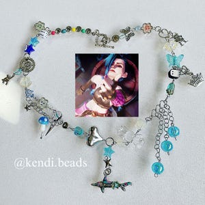 May include: A silver chain necklace with a variety of charms, including a heart, a mushroom, a star, a butterfly, a crown, and a shark. The necklace also features a small image of a character from the video game League of Legends. The necklace is made by @kendi.beads.
