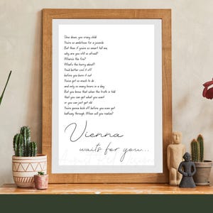 Op de afbeelding: Houten ingelijste print met de handgeschreven tekst van "Vienna". De print wordt weergegeven op een houten oppervlak met decoratieve items, waaronder een cactus, een klein beeldje en een vaas met rode bloemen. De tekst luidt "Vienna waits for you..."