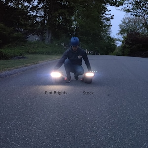 May include: A person crouches on a paved road, demonstrating the brightness of two sets of lights. The left light is labeled "Pint Brights" and the right light is labeled "Stock". The lights are bright white.