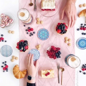 Puede incluir: Un mantel rosa con una variedad de aperitivos y bebidas, incluyendo pasteles, fruta y café. También hay posavasos con patrones azules y blancos en la mesa. La imagen incluye el texto "@Randlilieschen".