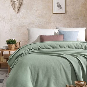 May include: A soft, sage green waffle weave throw blanket is folded on a bed with white and blue pillows. The blanket is made of a lightweight, breathable fabric that is perfect for year-round use.