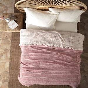 Könnte beinhalten: Draufsicht auf ein Bett mit weißen Kissen und einem gewebten Kopfteil. Eine rot-weiß gemusterte Decke mit Fransen bedeckt das Bett. Ein kleiner gewebter Beistelltisch hält eine Kerze und Karten. Das Bett steht auf einem braunen Teppich.