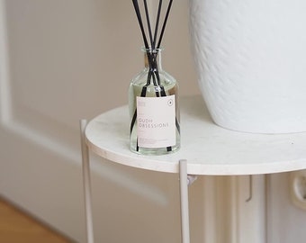 Peut inclure: Un diffuseur en verre transparent avec des bâtonnets noirs et une étiquette portant la mention "OUD OBSESSIONS" est posé sur une petite table ronde en marbre blanc. Un pot en céramique blanc avec une plante verte est en arrière-plan.