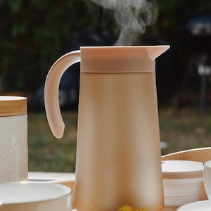 Könnte beinhalten: Eine beige Thermokanne mit einer breiten Öffnung und einem Griff. Dampf steigt aus der Kanne auf, was darauf hindeutet, dass sie mit einem heißen Getränk gefüllt ist. Die Kanne steht auf einem Holztablett mit einer weißen Tasse und Untertasse, einem kleinen weißen Behälter und ein paar gelben Blumen.