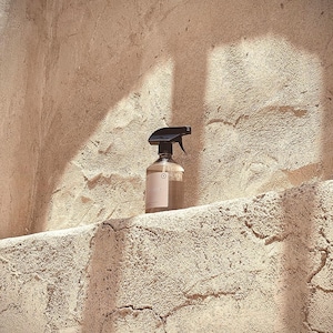 May include: A clear glass spray bottle with a black nozzle sits on a textured, light brown ledge. The bottle has a pink label with the word "RESOUE" and a circular design. The background is a light brown, textured wall.