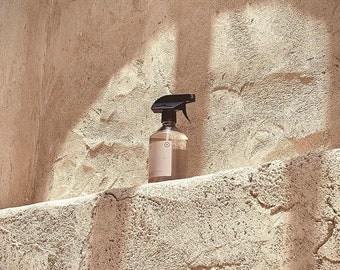 Peut inclure: Un flacon pulvérisateur en verre transparent avec une buse noire est posé sur un rebord texturé brun clair. La bouteille porte une étiquette rose avec le mot "RESOUE" et un motif circulaire. Le fond est un mur texturé brun clair.