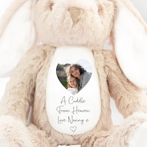 May include: A soft, beige bunny rabbit toy with long ears and a white heart-shaped patch on its chest. The patch displays a photo and the text "A Cuddle From Heaven Love Nanny x". The bunny is on a wooden surface.
