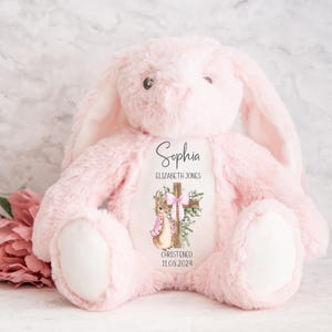 Pu&ograve; includere: Un coniglietto di peluche rosa con lunghe orecchie e zampe bianche. Il coniglietto ha un disegno stampato sul petto con una croce, un coniglio e il nome "Sophia" con la data "11.08.2024".
